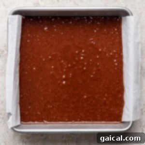 A rectangular metal baking pan lined with parchment paper is filled with s'mores brownies batter on a light textured surface.