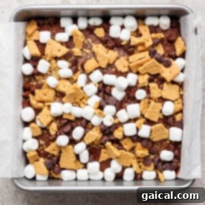 A square baking pan lined with parchment paper, filled with a mixture of mini marshmallows, chocolate chips, and broken graham crackers sits atop a rich s'mores brownies base.