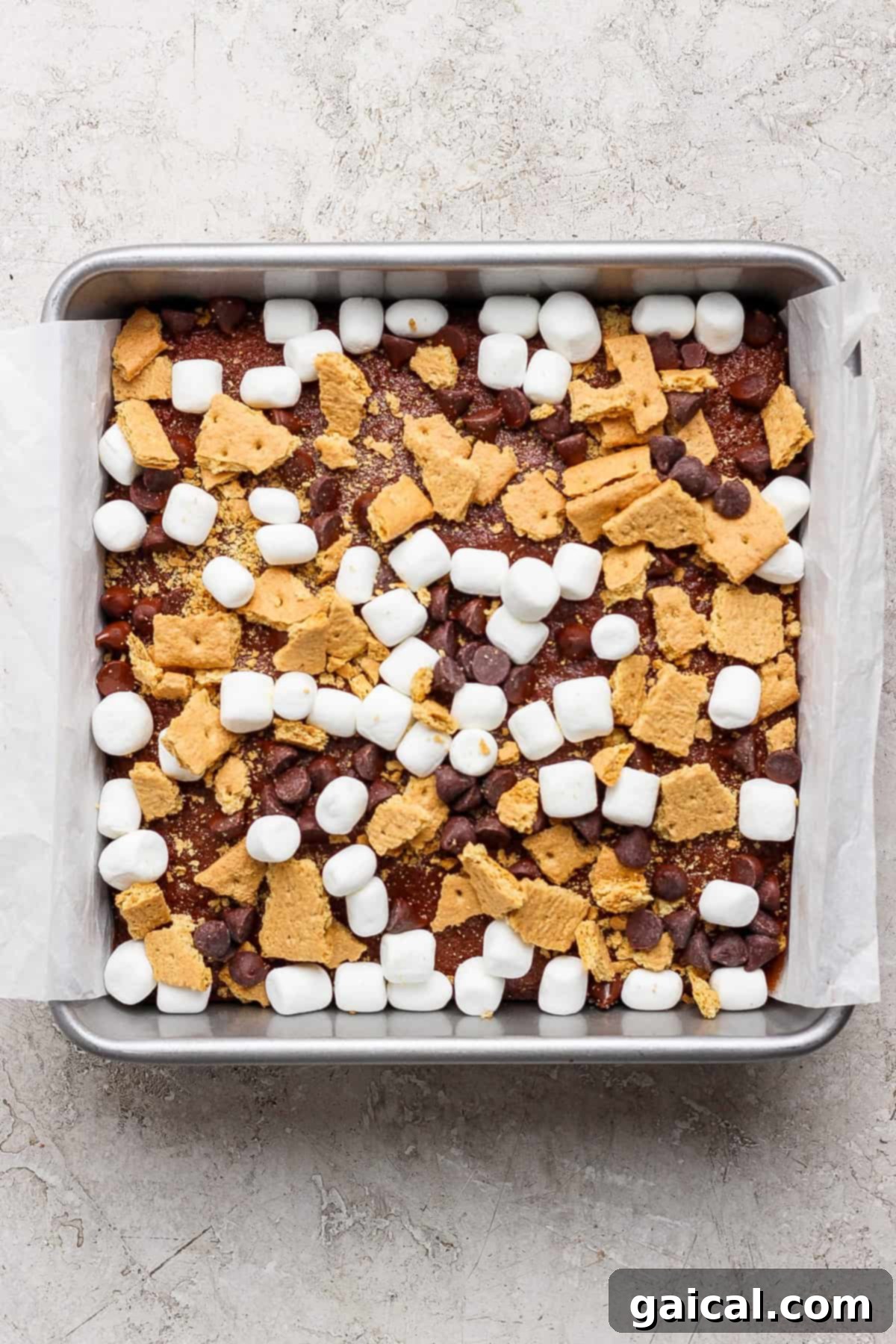 A square baking pan lined with parchment paper, filled with a mixture of mini marshmallows, chocolate chips, and broken graham crackers sits atop a rich s'mores brownies base.