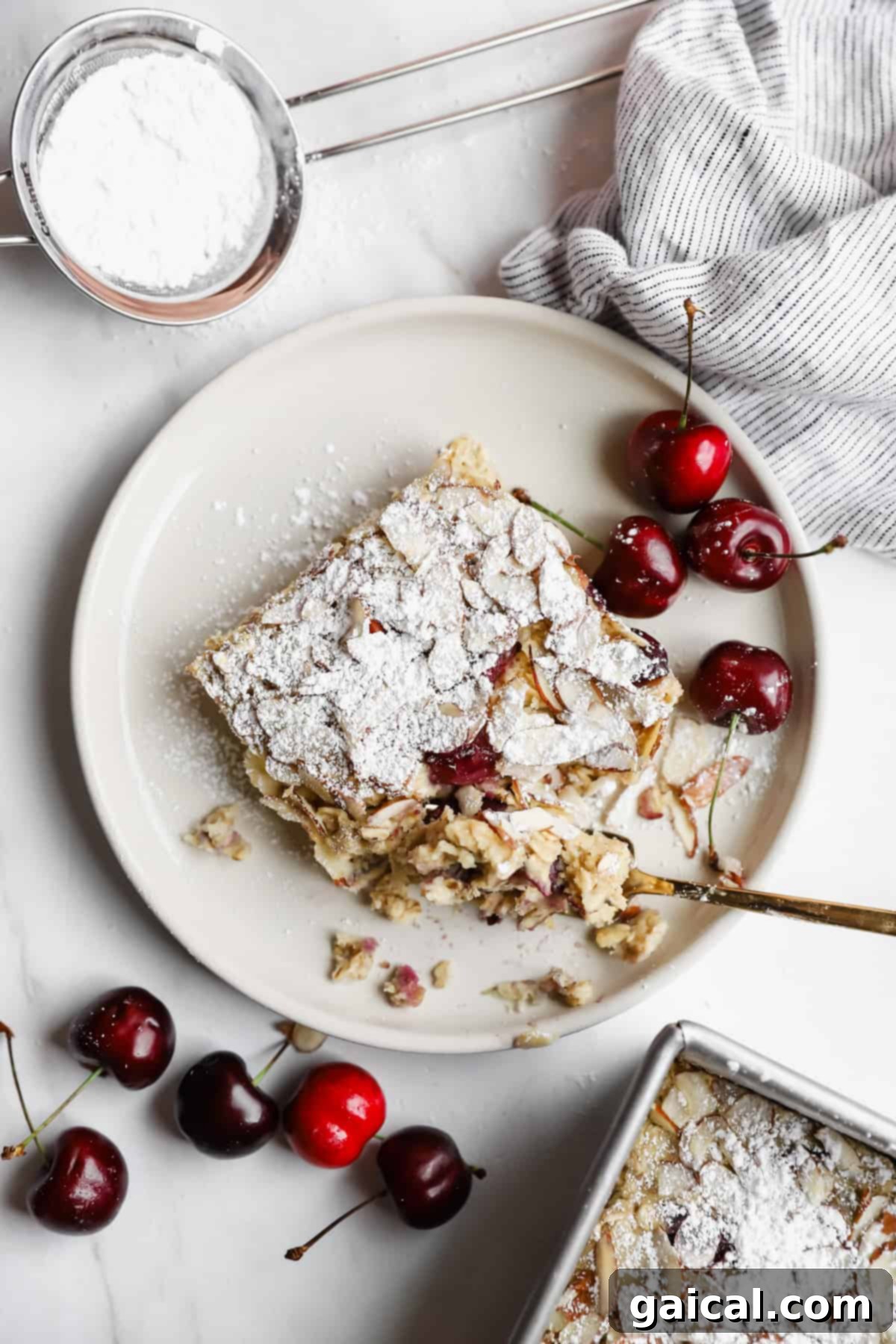 Bakery-Style Cherry Almond Baked Oats 2 A slice of cherry dessert dusted with powdered sugar on a plate, surrounded by fresh cherries, a striped cloth, and a sifter with powdered sugar.