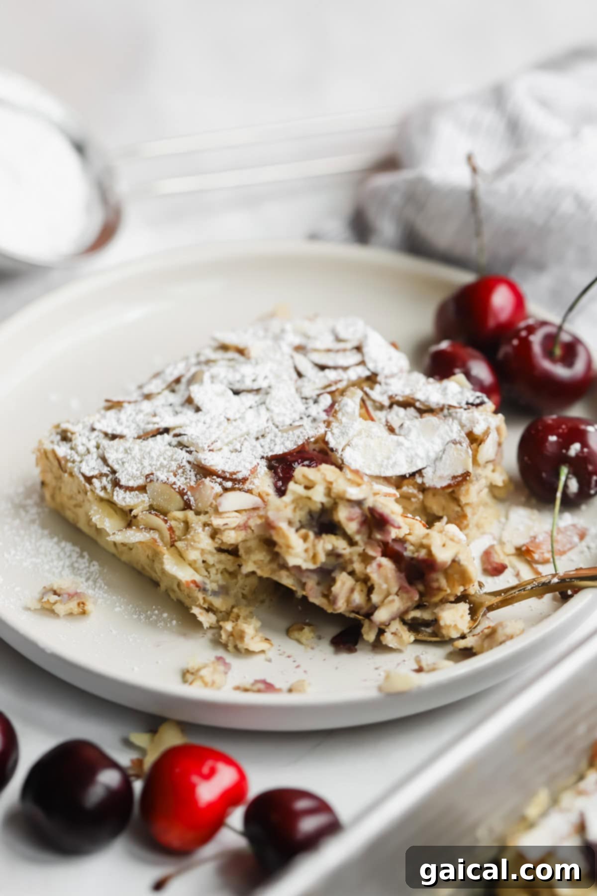Bakery-Style Cherry Almond Baked Oats 3 A partially eaten slice of almond and cherry cake topped with powdered sugar on a plate, with fresh cherries on the side.