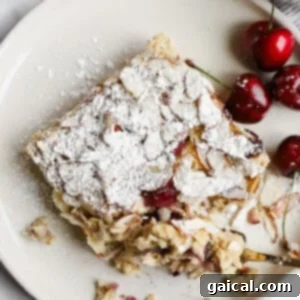 Bakery-Style Cherry Almond Baked Oats 4 A square slice of almond cherry cake topped with powdered sugar on a plate, served with fresh cherries on the side.