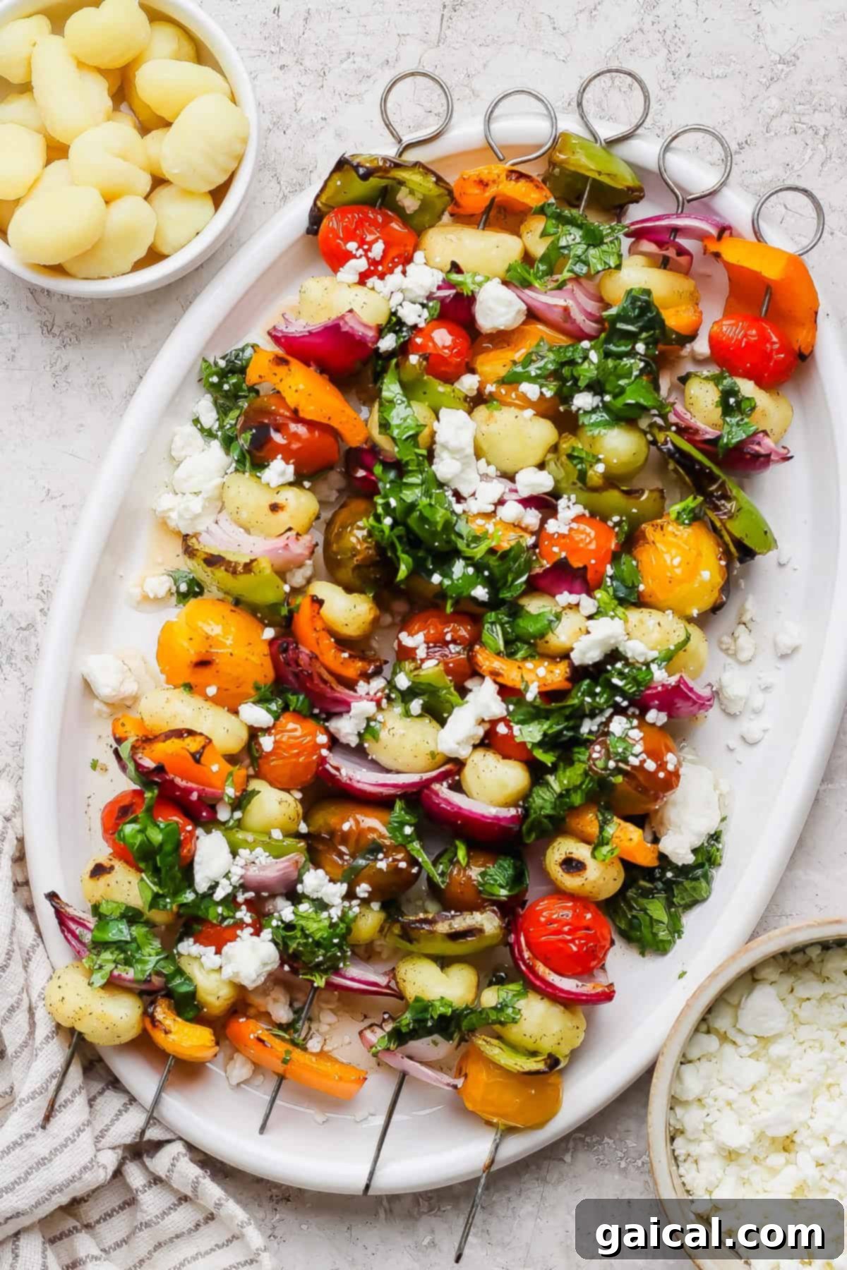 Vibrant Grilled Gnocchi Kebabs 2 A white platter with vegetable and gnocchi skewers, topped with chopped herbs and crumbled cheese, is pictured next to small bowls of cooked gnocchi and crumbled cheese.