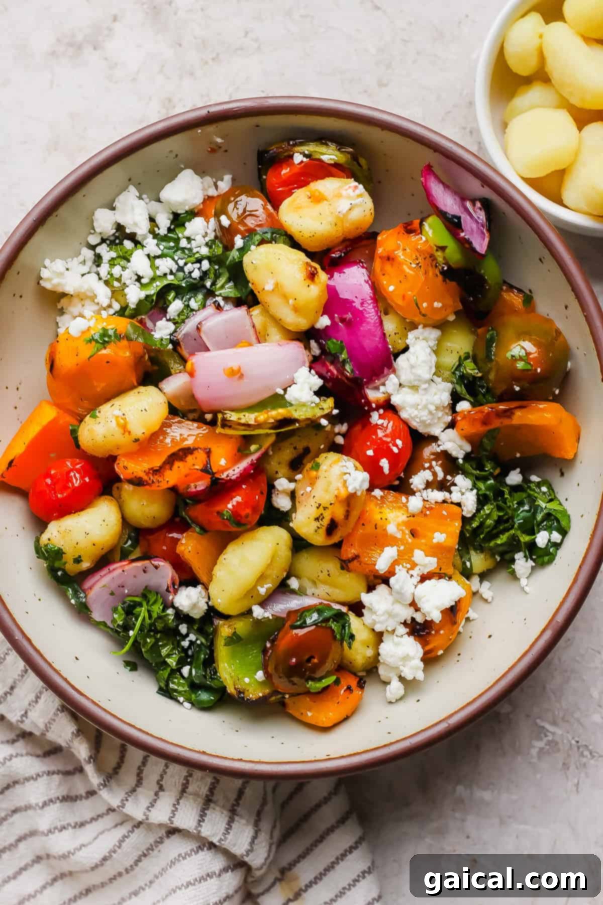 Vibrant Grilled Gnocchi Kebabs 4 A bowl of gnocchi mixed with roasted vegetables, red onion, tomatoes, leafy greens, and crumbled feta cheese, with a small bowl of gnocchi on the side.