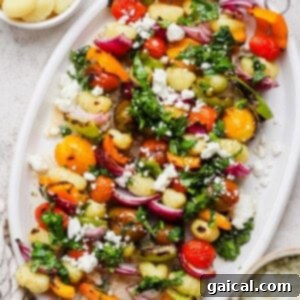 A white platter with gnocchi, roasted cherry tomatoes, bell peppers, red onions, fresh greens, and crumbled feta cheese.