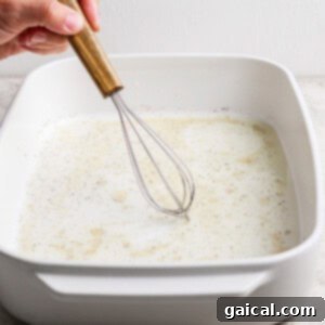 Vibrant Grilled Gnocchi Kebabs 7 A hand uses a wooden-handled whisk to mix a liquid with spices in a white rectangular baking dish on a light countertop.