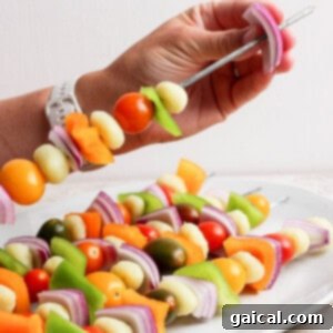 Vibrant Grilled Gnocchi Kebabs 8 A hand holds a metal skewer with pieces of onion, bell pepper, cherry tomatoes, and other vegetables above a plate of similar vegetable skewers.