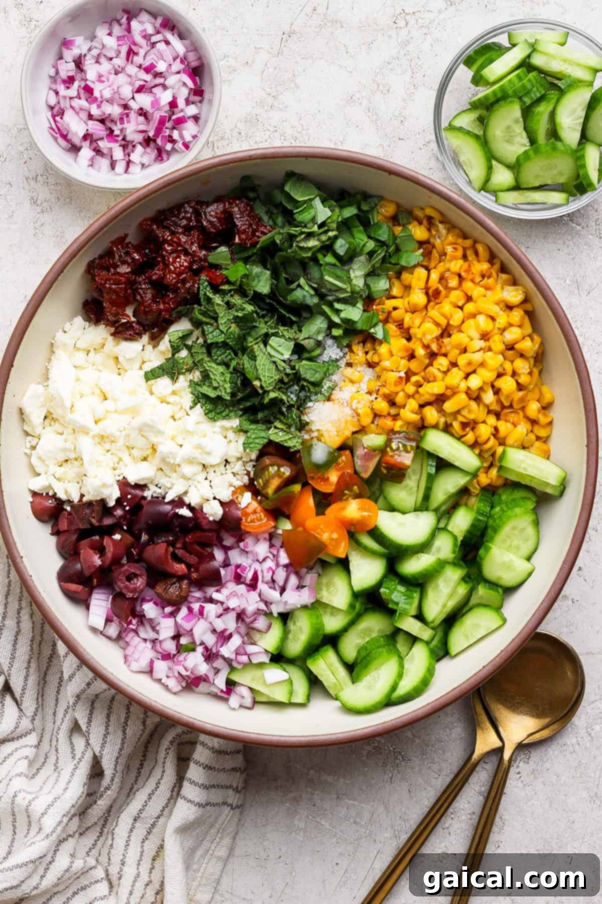 Mediterranean Golden Corn Salad 2 A large bowl containing chopped red onion, sun-dried tomatoes, feta cheese, olives, basil, mint, corn, cherry tomatoes, and cucumber, with bowls of onions and cucumbers on the side.