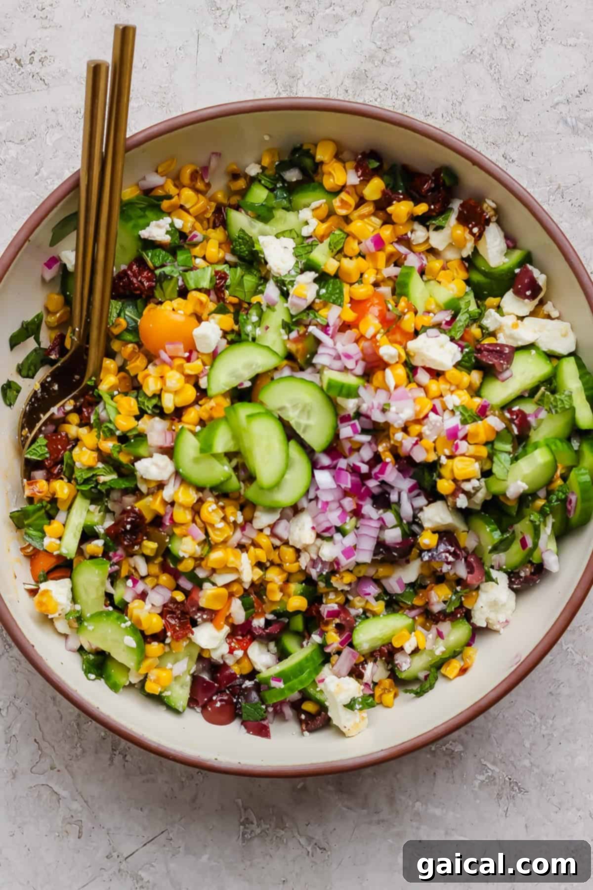 Mediterranean Golden Corn Salad 7 A bowl of Mediterranean corn salad with cucumbers, red onions, cherry tomatoes, feta cheese, leafy greens, and two gold forks.