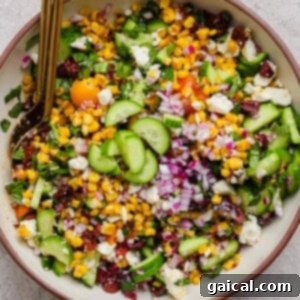 A bowl of chopped salad with corn, cucumbers, red onions, cherry tomatoes, feta cheese, and herbs, with a serving spoon.