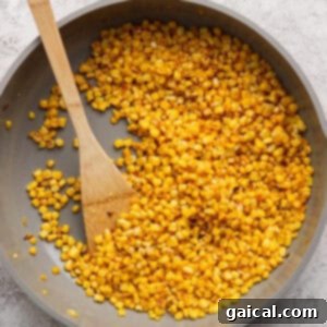 A skillet filled with sautéed corn kernels, perfect for a fresh Mediterranean corn salad, and a wooden spatula on a light gray surface.