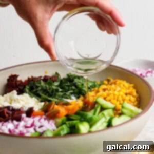 A hand pours dressing from a glass bowl onto a vibrant Mediterranean corn salad featuring cucumber, red onion, tomatoes, olives, feta cheese, and greens in a large white bowl.