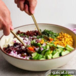A person prepares a mediterranean corn salad, mixing chopped cucumber, corn, red onion, olives, sun-dried tomatoes, greens, and feta cheese in a large bowl using two gold utensils.
