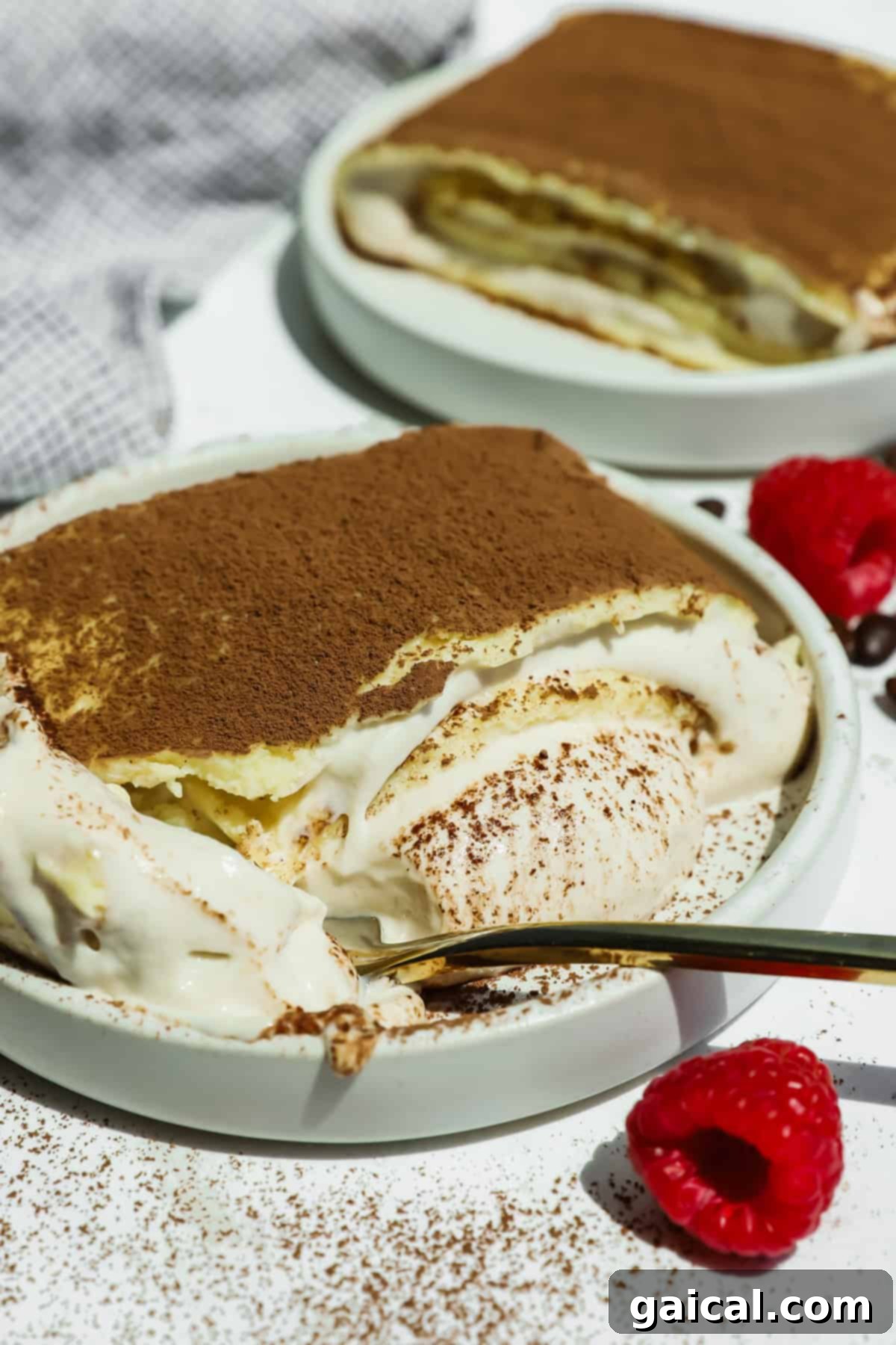 A beautifully presented slice of cottage cheese tiramisu crepe roll with creamy layers and a generous dusting of cocoa powder. Fresh raspberries and a gold spoon are artfully placed beside it.