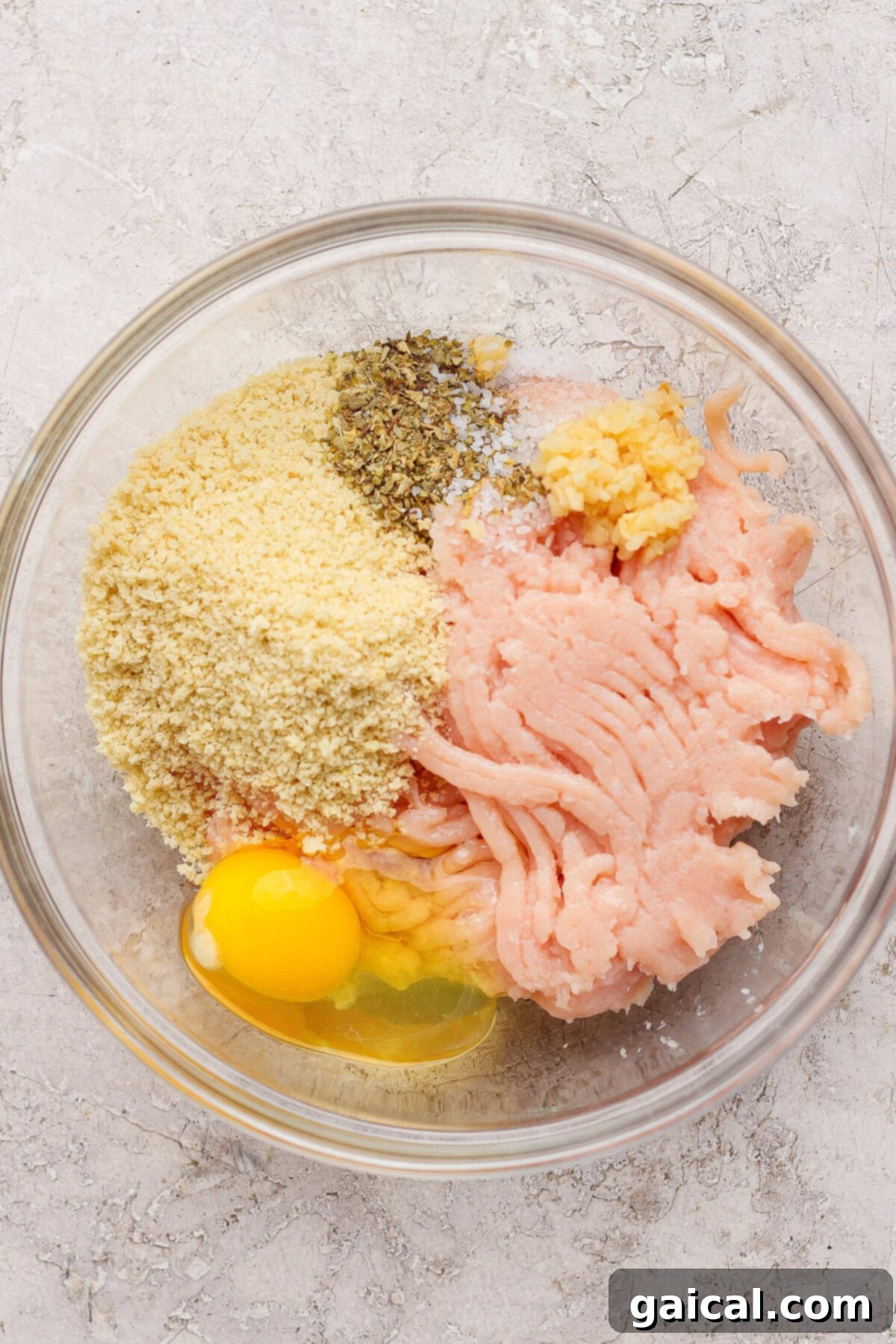 Easy Summer Skillet Orzo with Zucchini and Chicken Meatballs 3 A glass bowl containing ground chicken, a cracked egg, breadcrumbs, minced garlic, salt, and dried herbs on a light textured surface.