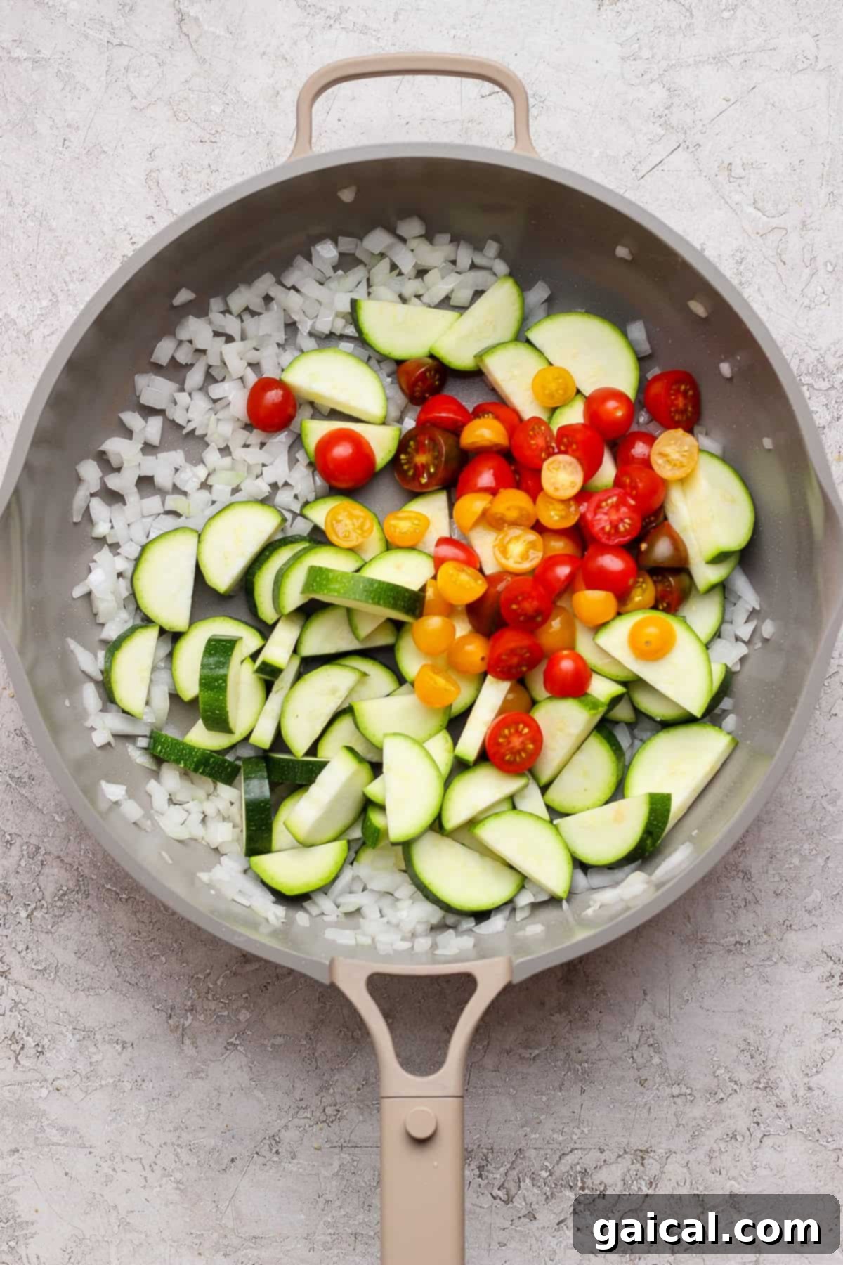 Easy Summer Skillet Orzo with Zucchini and Chicken Meatballs 5 A frying pan contains chopped onions, sliced zucchini, and halved cherry tomatoes on a light textured surface.
