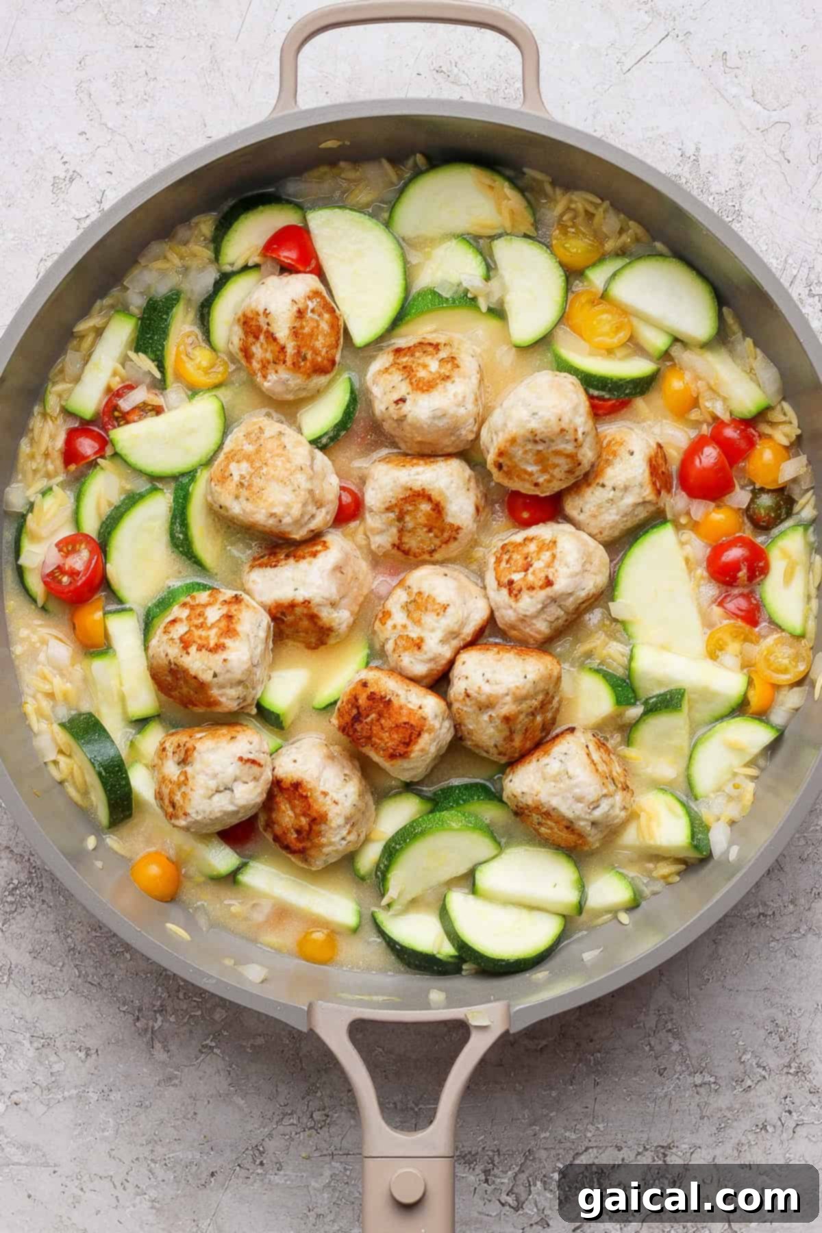 Easy Summer Skillet Orzo with Zucchini and Chicken Meatballs 6 A skillet with browned meatballs, sliced zucchini, yellow squash, cherry tomatoes, and orzo in a light sauce on a gray surface.