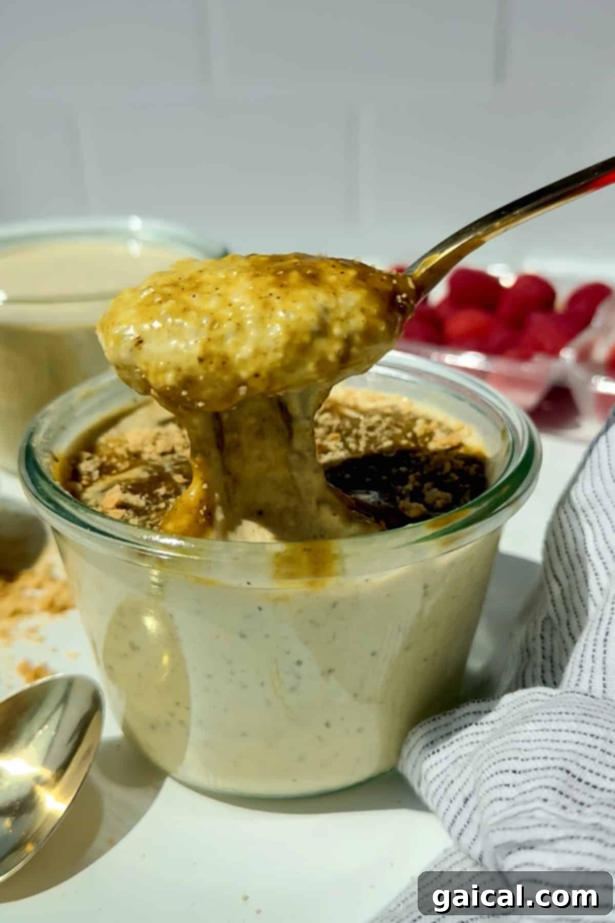 A spoon lifts creamy overnight oats from a glass jar, topped with a drizzle of syrup and a sprinkle of crumbs, with raspberries and a striped cloth nearby. The scene suggests a fresh and delicious breakfast.