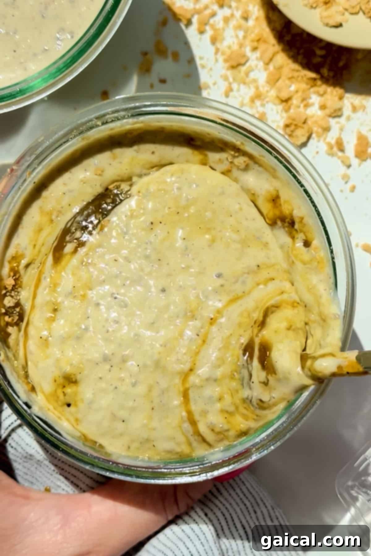 A glass bowl containing a creamy, beige mixture with swirls and specks, being stirred with a spoon. Crumbled pieces are scattered nearby on a white surface, highlighting the rich texture of the oats.