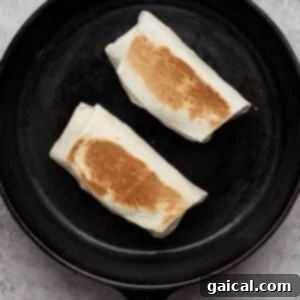 Two folded burritos being toasted in a black cast iron skillet on a light textured surface, achieving a golden brown crisp.