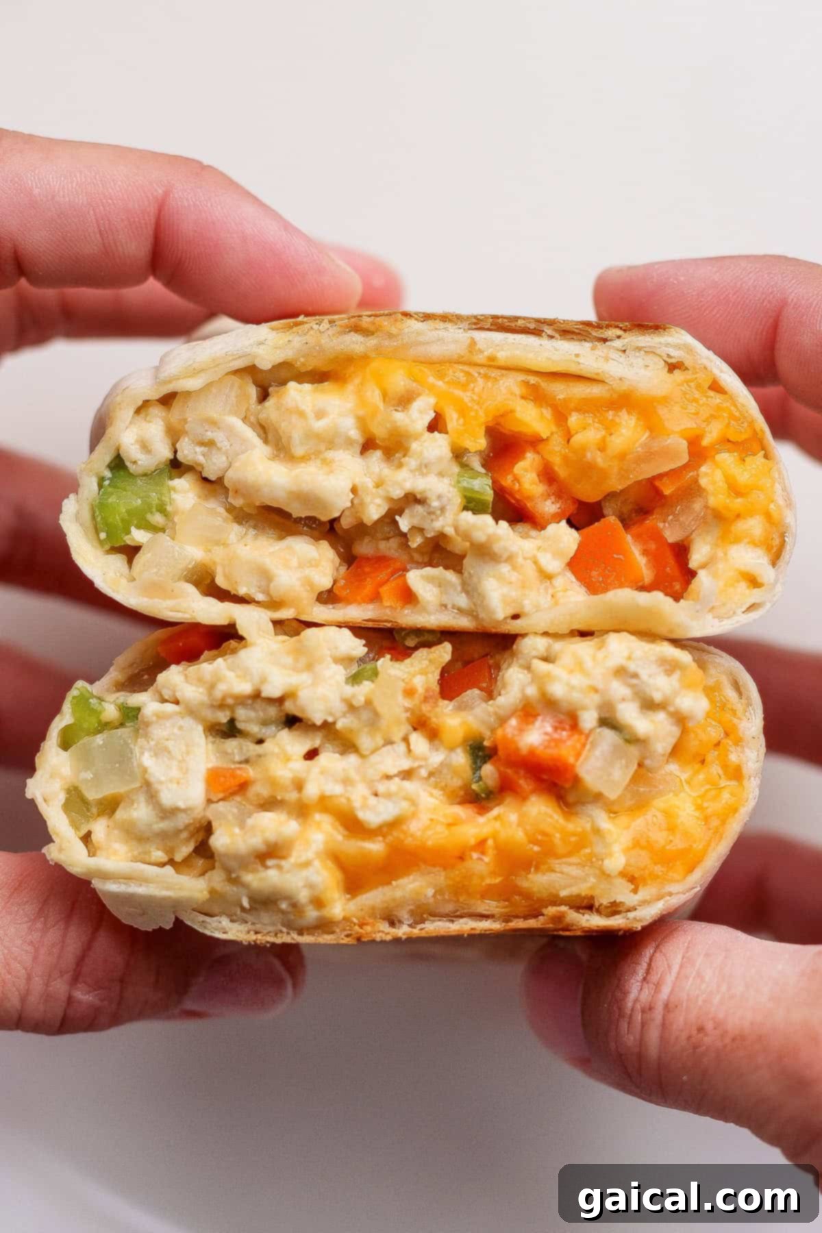 Protein Packed Buffalo Chicken Burritos 4 Two hands hold a breakfast burrito cut in half, showcasing the rich filling of scrambled eggs, melted cheddar cheese, and colorful diced vegetables inside a warm tortilla wrap.