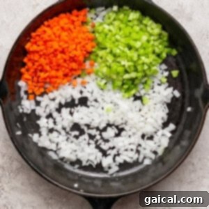 A cast iron skillet containing diced carrots, celery, and onions arranged in separate sections on a light textured surface, ready to be cooked.