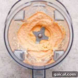 A blender with creamy, light orange mixture spread around the blades, viewed from above on a textured surface, showcasing the smooth texture of the buffalo sauce.