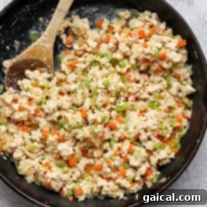 A cast iron skillet filled with scrambled tofu mixed with diced carrots, celery, onions, and a wooden spoon on the side, representing the buffalo chicken mixture.