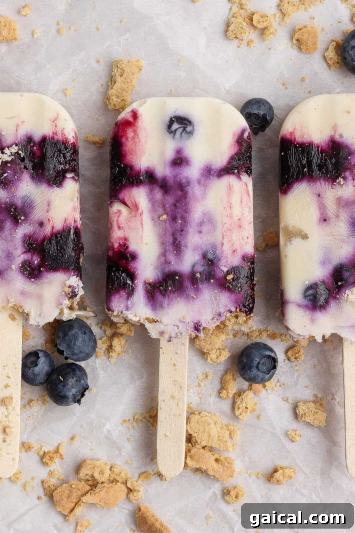 Three blueberry cheesecake popsicles with visible blueberry swirls are arranged on a surface scattered with fresh blueberries and crumbled graham crackers.