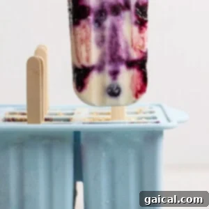 A hand holds a popsicle with purple, white, and red swirls above a blue silicone popsicle mold containing more popsicles.