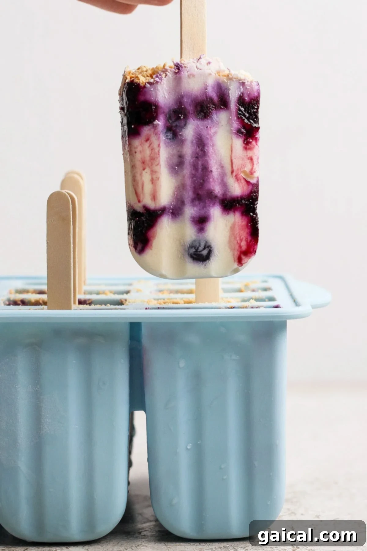 A hand holds a popsicle with purple, white, and red swirls above a blue silicone popsicle mold containing more popsicles.