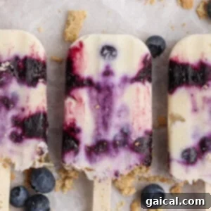 Three creamy blueberry popsicles with purple swirls are arranged on a light surface, surrounded by scattered blueberries and crumbled cookie pieces.