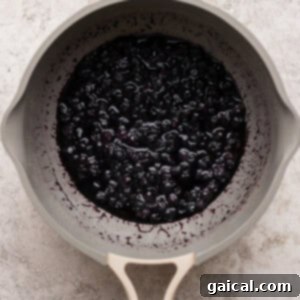 A beige saucepan filled with cooked blueberries in a thick purple sauce on a light textured surface.