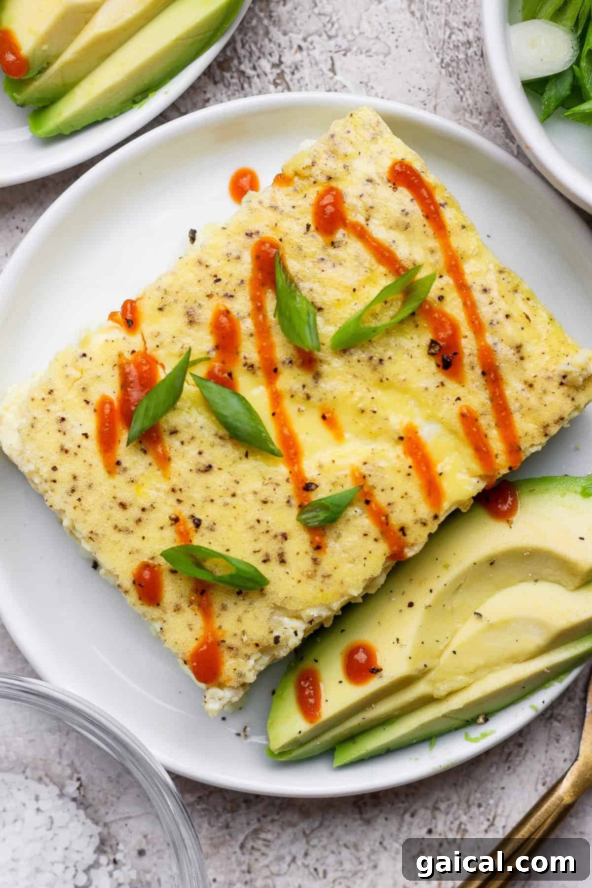 A rectangular egg dish topped with sliced green onions and drizzled with hot sauce, served with avocado slices on a white plate.