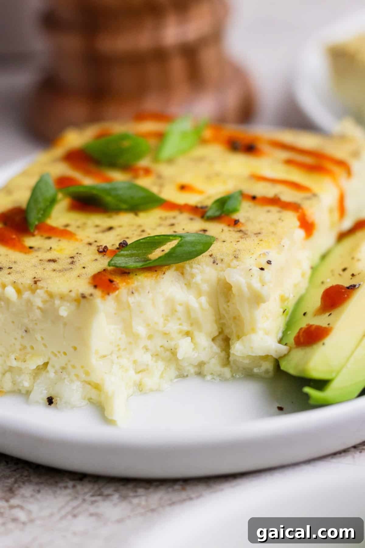 A slice of baked egg casserole topped with sliced green onions and hot sauce, served with sliced avocado on a white plate.