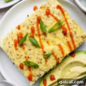 A rectangular egg dish garnished with sliced green onions and hot sauce, served with avocado slices on a white plate.