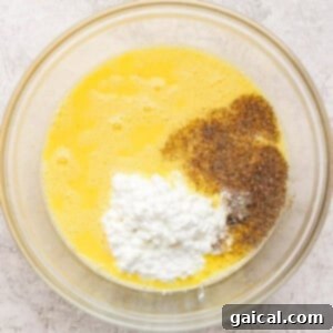 A glass bowl containing whisked eggs, cottage cheese, ground black pepper, and salt on a light textured surface.