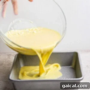 A hand pours yellow cake batter from a glass bowl into a square metal baking pan on a countertop.