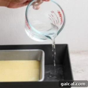 A hand pours water from a glass measuring cup into a larger baking pan, which holds a smaller pan filled with batter.
