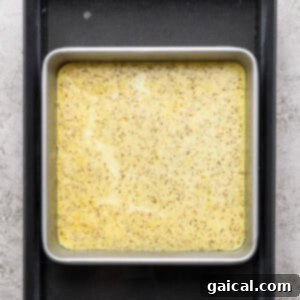 A square metal baking pan filled with a yellow speckled batter sits inside a larger black tray on a textured gray surface.