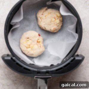 Two unbaked dough rounds with visible apple chunks, placed on parchment paper inside an air fryer basket, ready to be cooked.