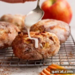A hand drizzles white icing from a spoon onto a warm, round baked apple fritter with visible apple pieces, resting on a cooling rack.