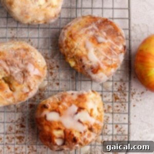 Four perfectly glazed apple fritters sit on a cooling rack, surrounded by fresh apples, cinnamon sticks, and apple slices on a light surface, ready to be enjoyed.