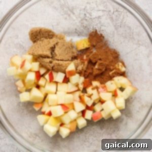 A glass bowl containing diced apples, brown sugar, and ground cinnamon on a light textured surface, ready for mixing.