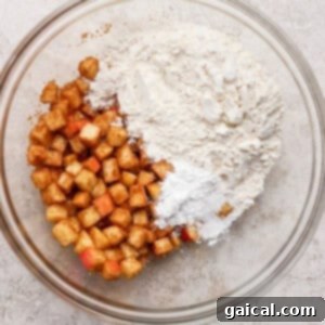 A glass bowl with diced apples coated in cinnamon on one side and a mixture of flour and white powder (baking powder/salt) on the other, on a light textured surface.