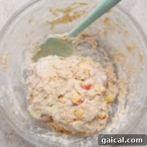 A glass bowl containing dough mixed with small apple chunks and a green spatula, showing the dough forming, on a light textured surface.