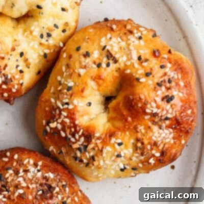 A delightful close-up of three perfectly golden-brown bagels, generously adorned with savory sesame seeds and other classic seasonings, artfully arranged on a soft beige plate, eagerly awaiting to be sliced and enjoyed.