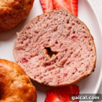 A beautifully sliced strawberry bagel is artfully presented on a clean white plate, with two fresh, vibrant strawberry slices delicately placed underneath, creating an inviting and refreshing culinary display.