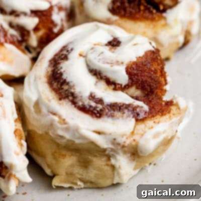 A close-up of a delectable cinnamon roll, generously adorned with creamy white icing and intricate swirls of cinnamon sugar, creating an inviting and comforting culinary image on a light-colored plate.