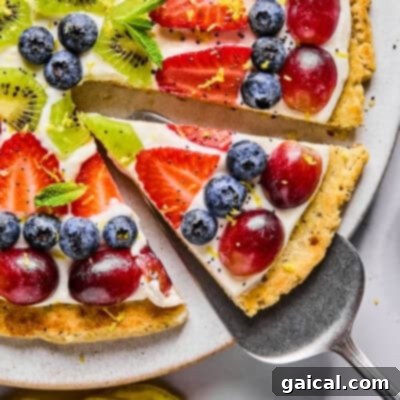 A vibrant and refreshing slice of fruit pizza, beautifully adorned with an assortment of fresh blueberries, succulent strawberries, juicy grapes, bright kiwi slices, and a delicate sprinkle of mint leaves. This colorful dessert rests on a wholesome cottage cheese cookie crust, generously spread with a luscious cream cheese frosting, making it a perfect treat.